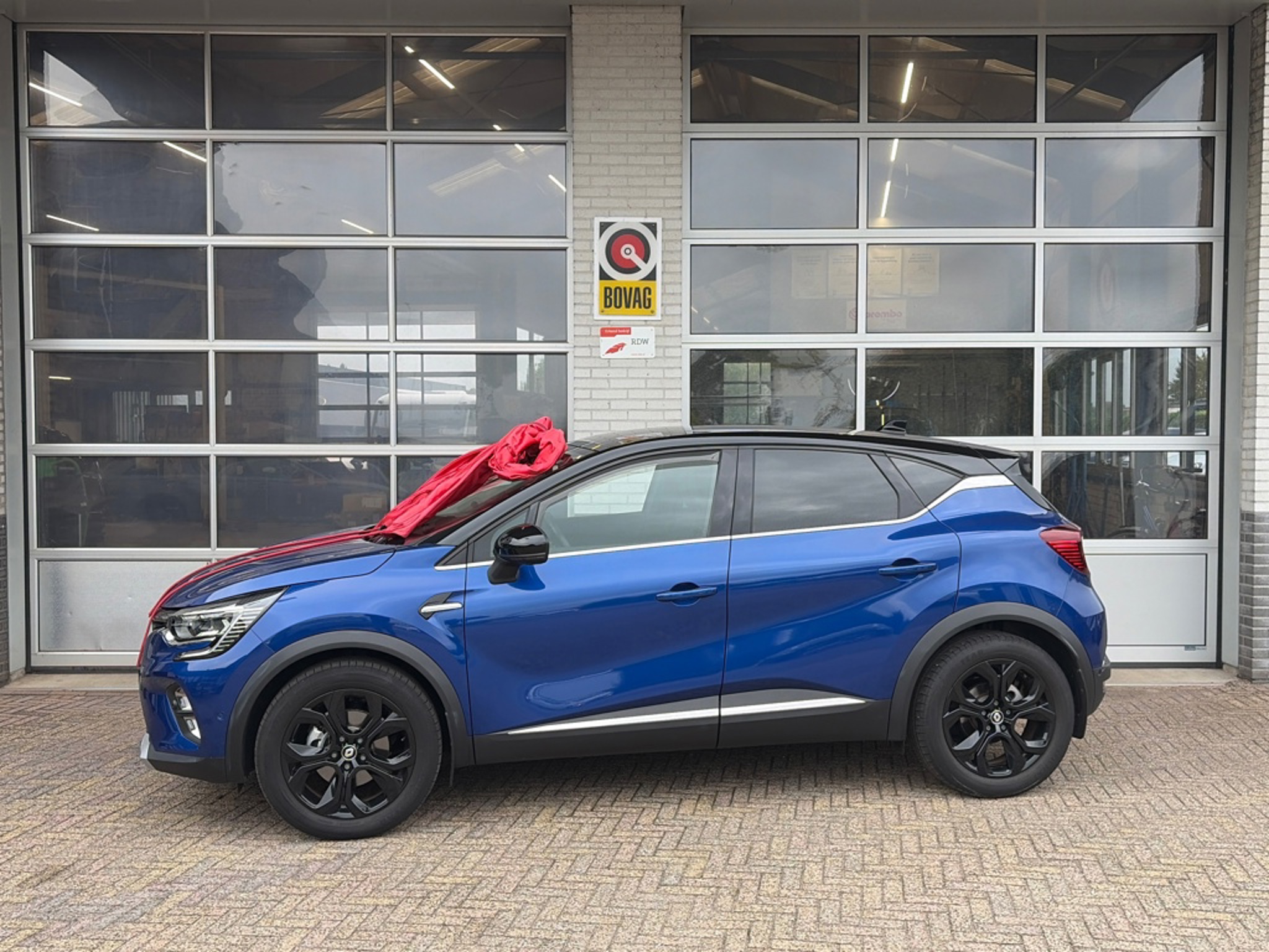 Blauwe Renault Captur met een rode afleverstrik voor de glazen werkplaatsdeuren van Autobedrijf Van Oostrum, met het BOVAG-bord aan de gevel